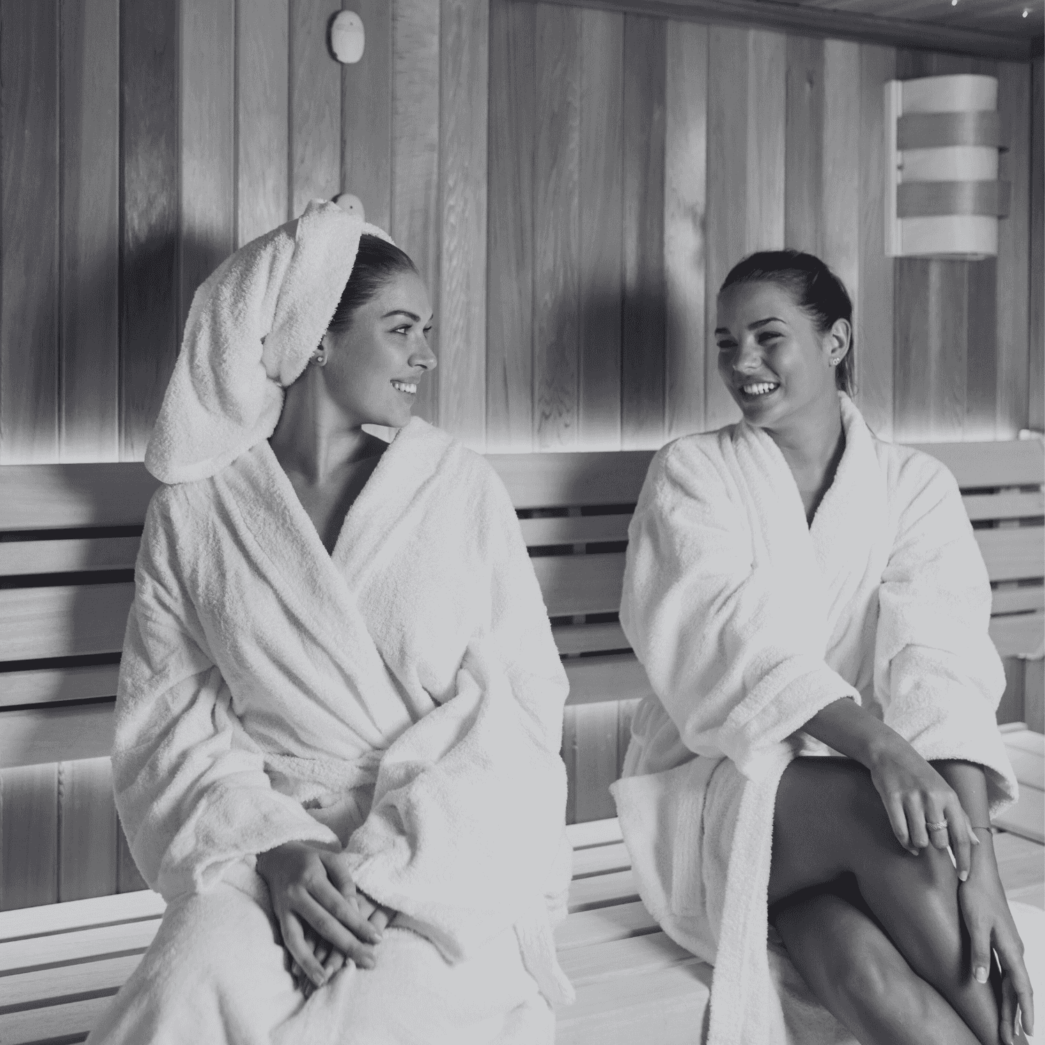Two women in bathrobes smiling and sitting inside a wooden sauna.